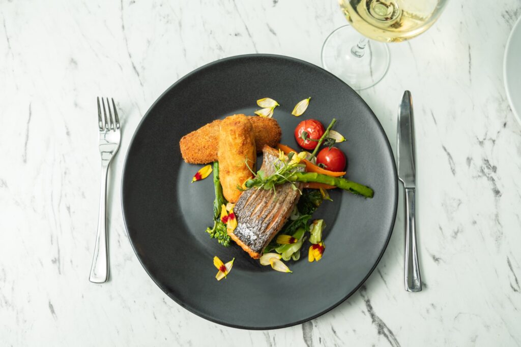 Elegant seared fish with asparagus, cherry tomatoes, and crispy potatoes, paired with white wine on a marble table.