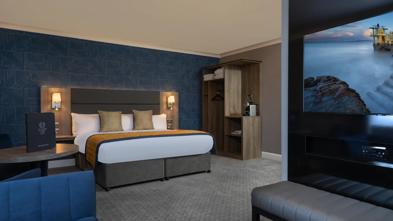 Elegant hotel room with plush king bed, stylish navy decor, modern TV, and open wardrobe, promising comfort and relaxation.