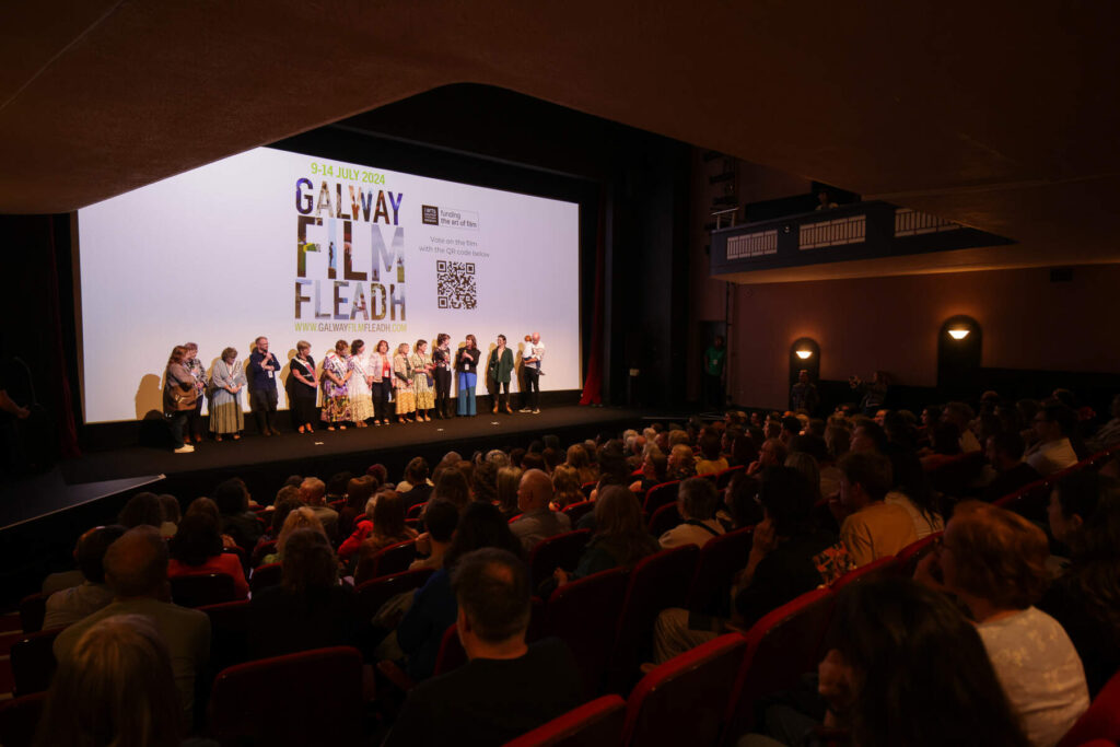 People in a dark room stand before a stage that has a white backdrop with Galway Film Fleadh projected onto it.