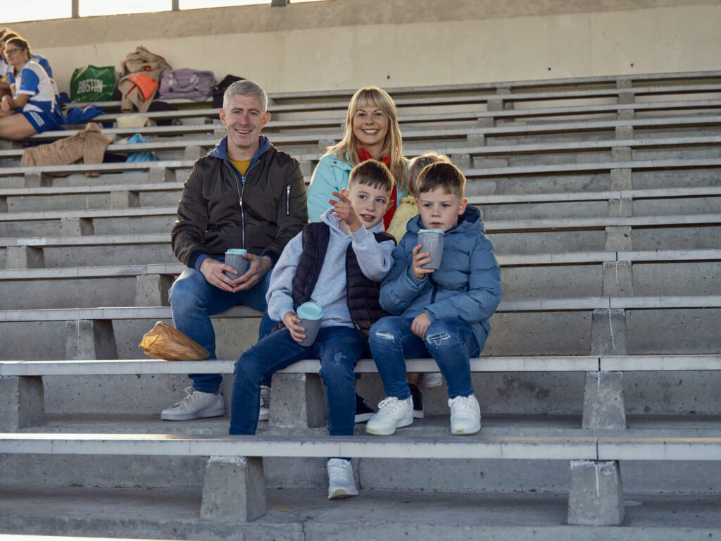 A Happy family sit in a stadium looking at the GAA game.