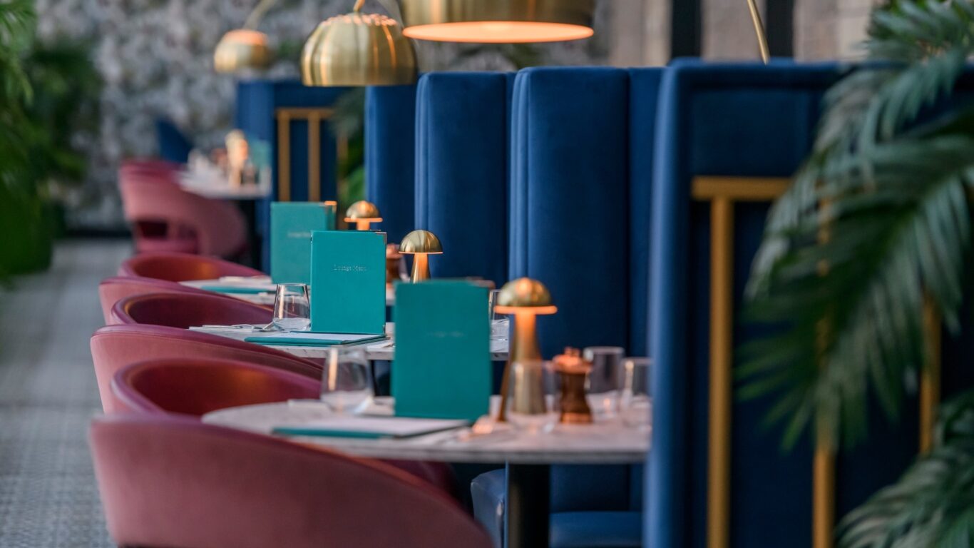 Elegant hotel dining area with plush pink chairs, blue booths, golden lamps, leafy plants, creating a cosy atmosphere.
