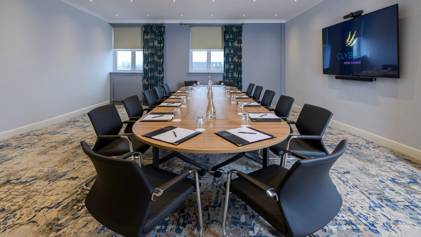 Elegant meeting room with a large oval table, modern chairs, natural light, and a flat-screen TV for presentations.