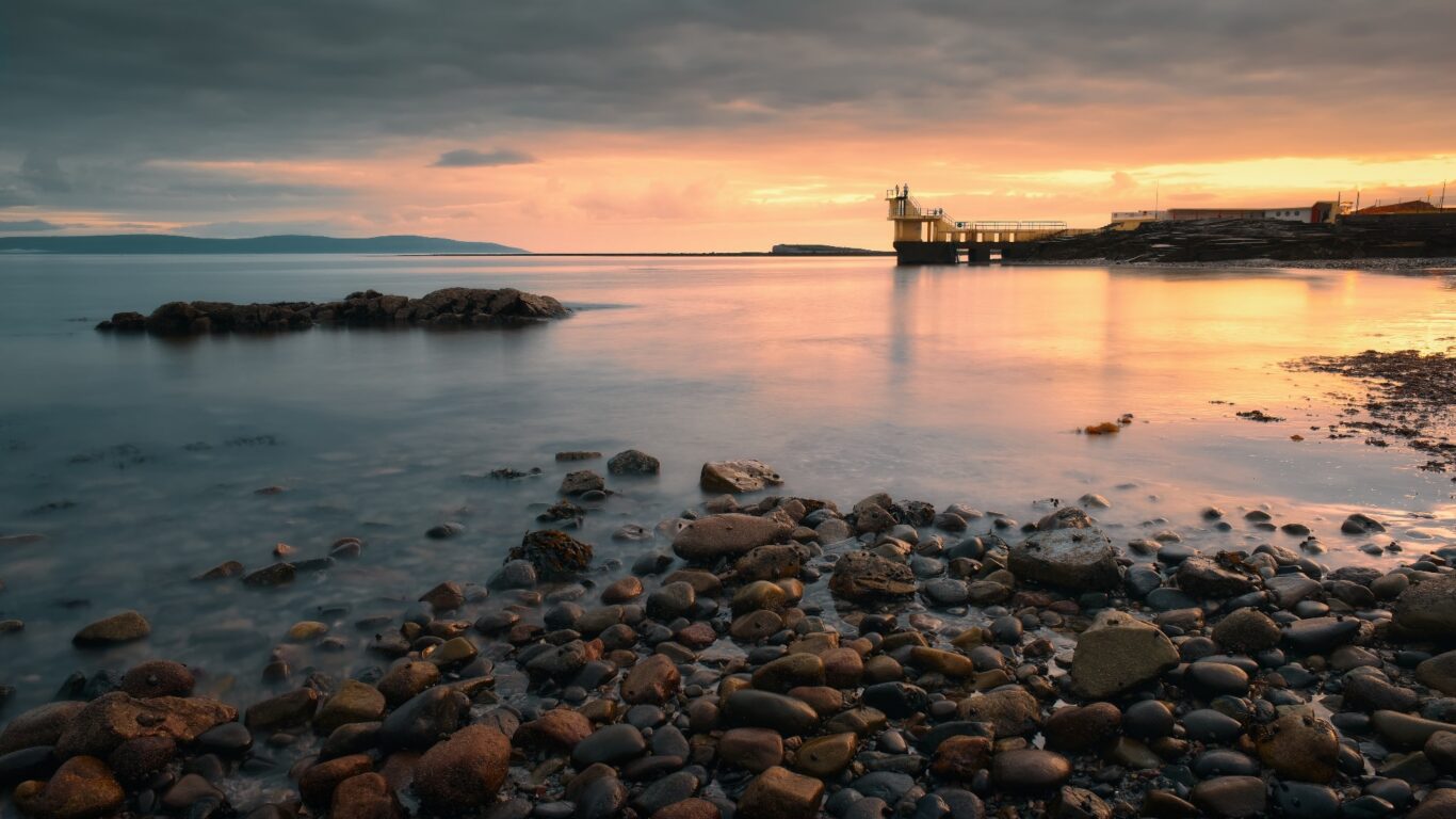 Tranquil coastal sunset view from rocky shore, highlighting serene waters and a distant diving tower. Perfect for relaxation.