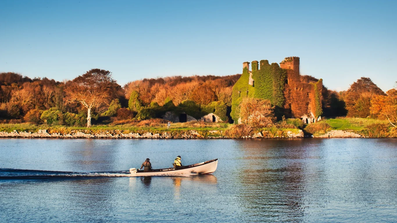 Two people in a small boat travel on a calm river, passing by the ivy-covered ruins of an old castle surrounded by autumn trees.