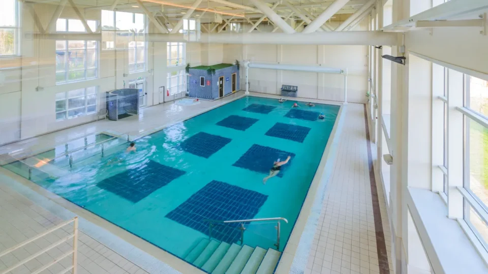 Indoor swimming pool with a few people swimming, large windows on one side, blue tiles on the pool floor, and a stairway leading into the water.