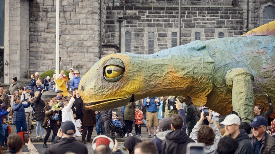 A large, colorful lizard puppet is paraded through a crowd of people in front of a stone building. Many onlookers are taking photos and watching the event.