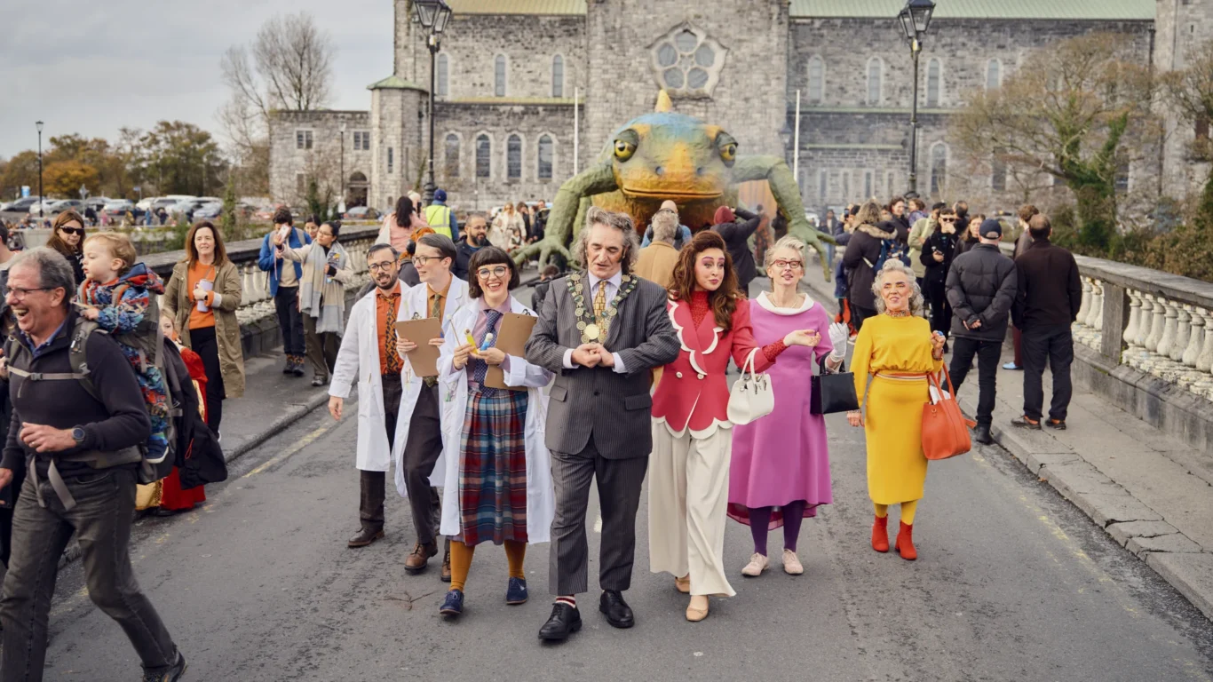 A group of people in colorful costumes walk down a street, led by a man in a chain of office, with a large frog puppet and a stone building in the background.