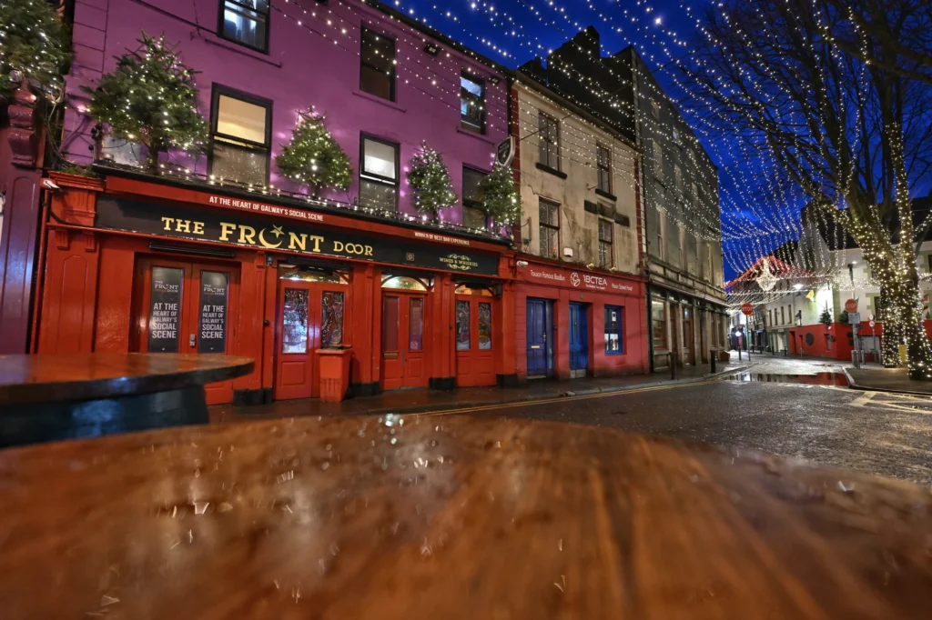 A brightly lit street at night with decorated trees, string lights overhead, and a red pub called "The Front Door" on the left—capturing the festive spirit of the Galway Christmas Market.