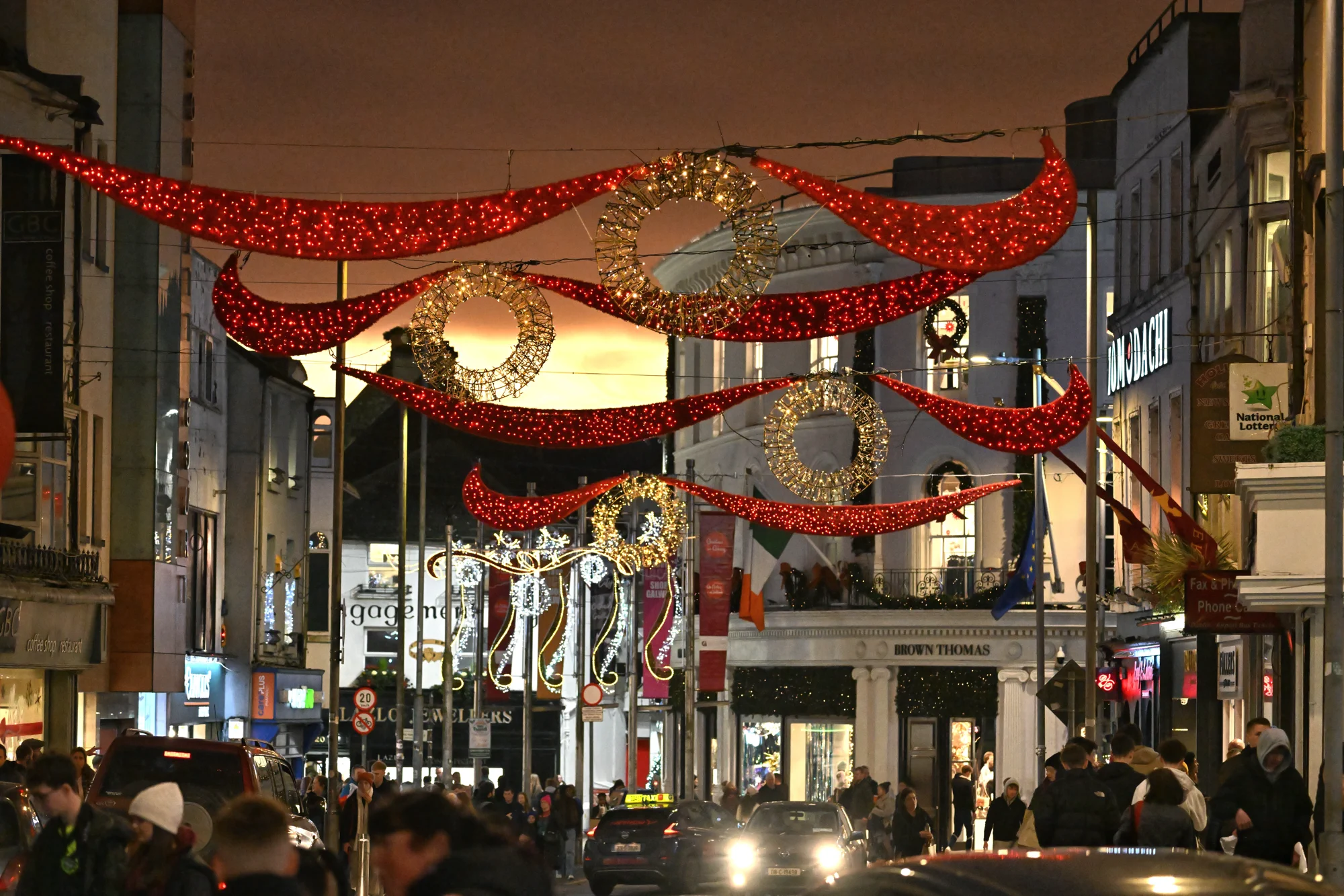 A busy city street at dusk, reminiscent of the Galway Christmas Market, is decorated with red and gold holiday lights and ornaments, with people walking and cars driving below.