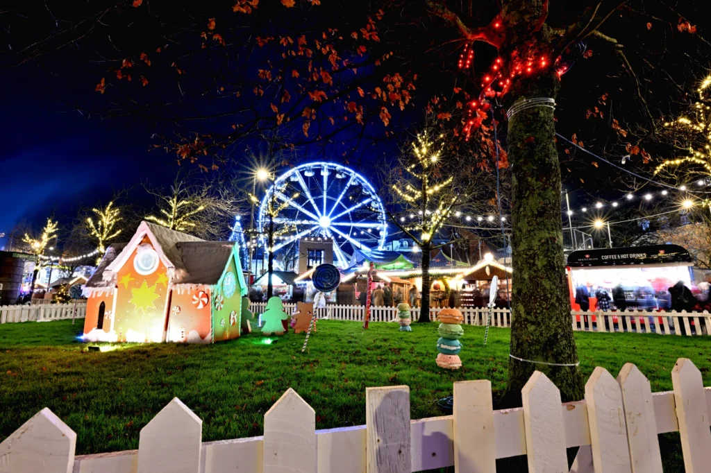 A night scene of the Galway Christmas Market with a festive fair, Ferris wheel, decorated gingerbread house, string lights, and a white picket fence in the foreground.