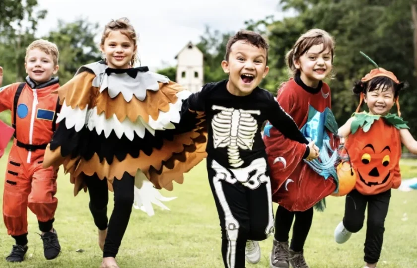 Five children dressed in various colorful costumes, including a skeleton, pumpkin, and bird, run together on a grassy field outdoors, smiling and holding hands.