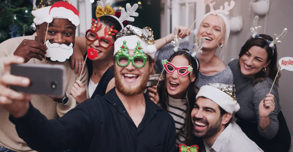A group of seven people wearing festive holiday costumes and accessories smile and pose for a selfie together at a party.