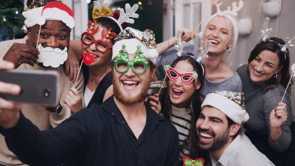 A group of seven people wearing festive holiday costumes and accessories smile and pose for a selfie together at a party.