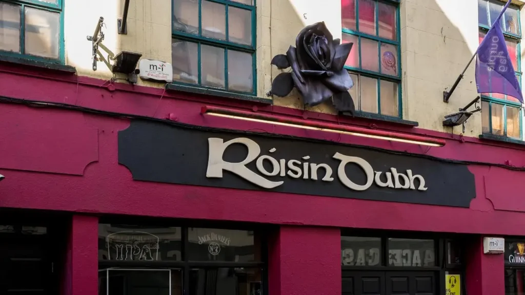 A building facade with a sign reading "Róisín Dubh" in stylized letters, featuring a large black rose sculpture above the entrance, often decorated for events like the Halloween parade in Galway.