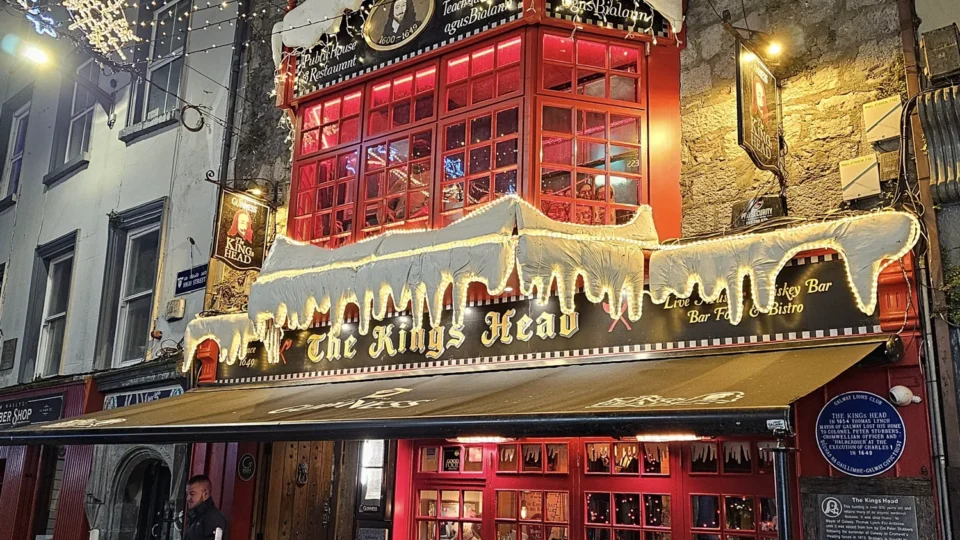 Street view of The King's Head pub with red window frames, festive lights, artificial snow decorations, and empty outdoor seating at night.