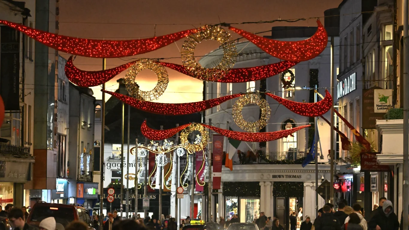 A busy city street at dusk decorated with red and gold holiday lights, with people walking and cars driving below the festive displays.