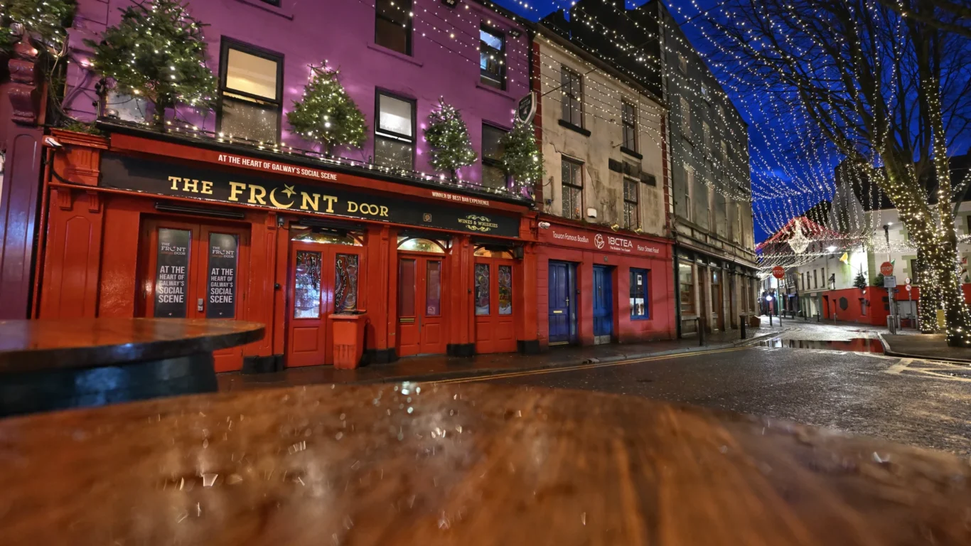 A street view at night shows The Front Door pub with red doors, holiday lights, and decorated trees on its facade; nearby buildings and string lights are visible overhead.