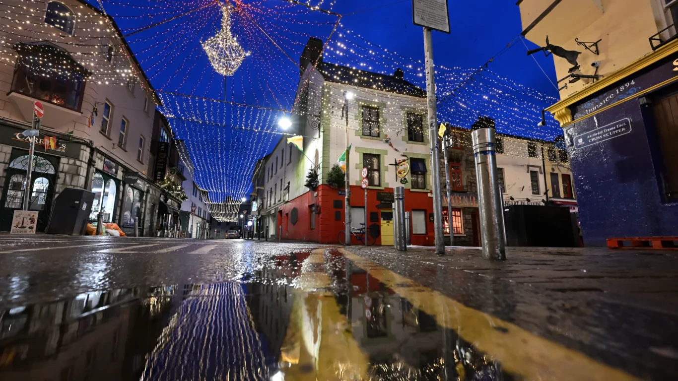 A cobblestone street with festive string lights overhead is reflected in a puddle at night, surrounded by buildings and shopfronts.