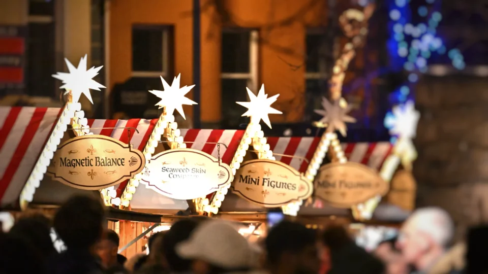 Crowd gathers in front of festive market stalls with striped roofs and illuminated star decorations; signs read