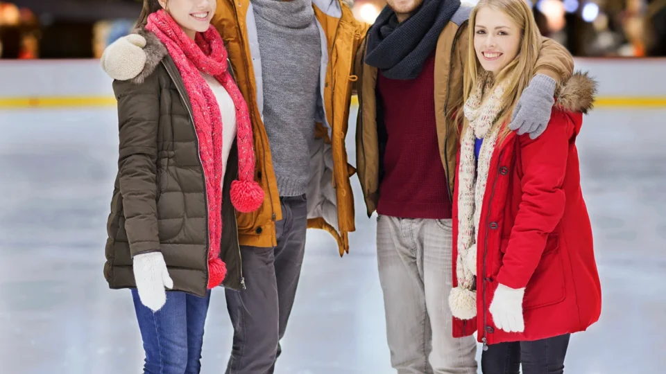 Four people in winter clothing are standing together on an outdoor ice skating rink with a lit Christmas tree and string lights in the background.