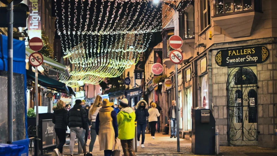 People walk along a cobblestone street decorated with string lights at night; shops and a sweater shop are visible on both sides of the street.