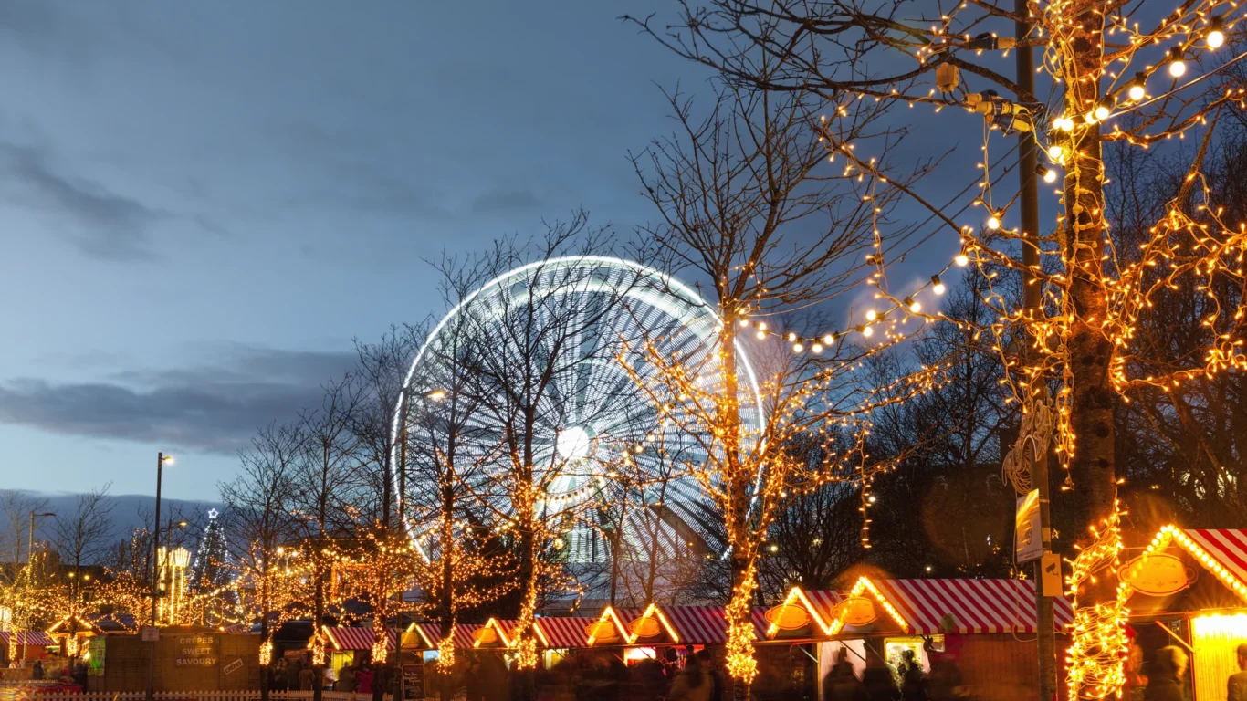 Festive market stalls with string lights and a large illuminated Ferris wheel in the background at dusk, with trees and some people visible.
