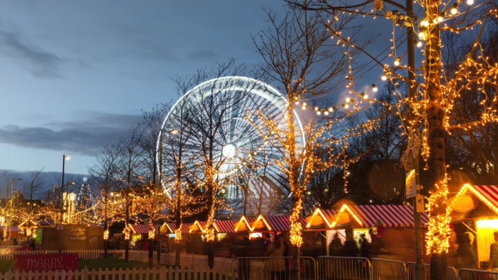 Festive market stalls with string lights and a large illuminated Ferris wheel in the background at dusk, with trees and some people visible.