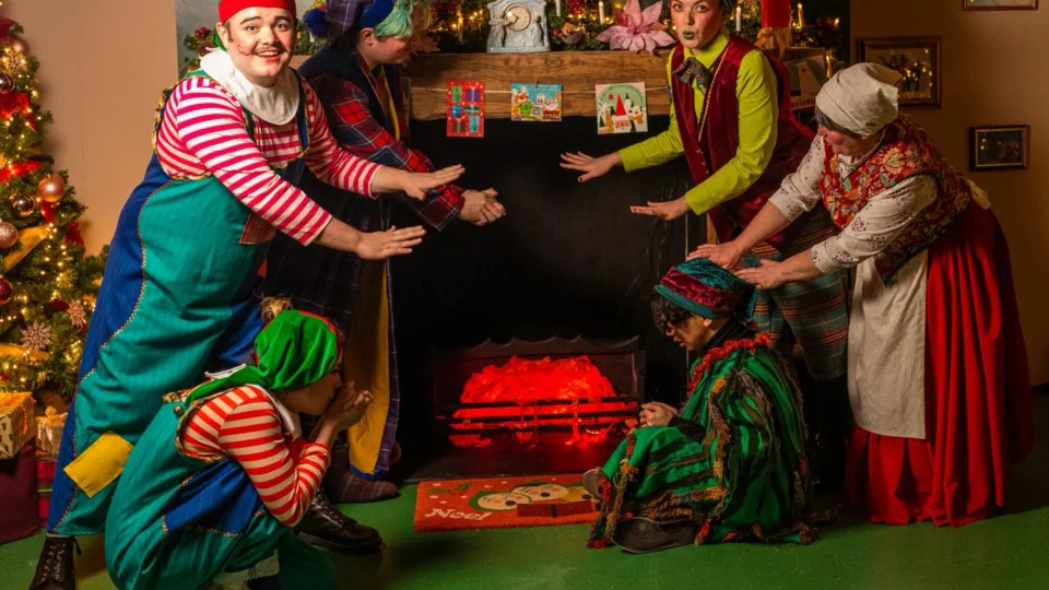 Six people in colorful holiday costumes pose around a decorated faux fireplace, with a lit Christmas tree and festive decorations in the background.