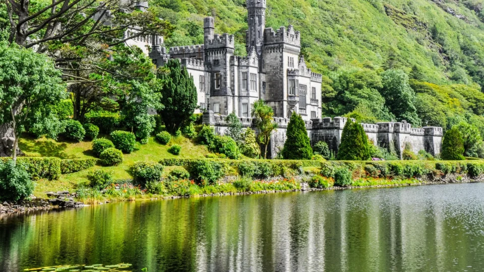 A large stone castle with towers and battlements stands beside a reflective lake, surrounded by lush greenery and forested hills.