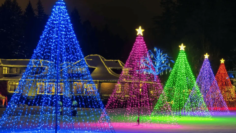 Four large, cone-shaped Christmas light displays in blue, pink, green, and red with star toppers stand on a snowy lawn in front of a house decorated with string lights at night.