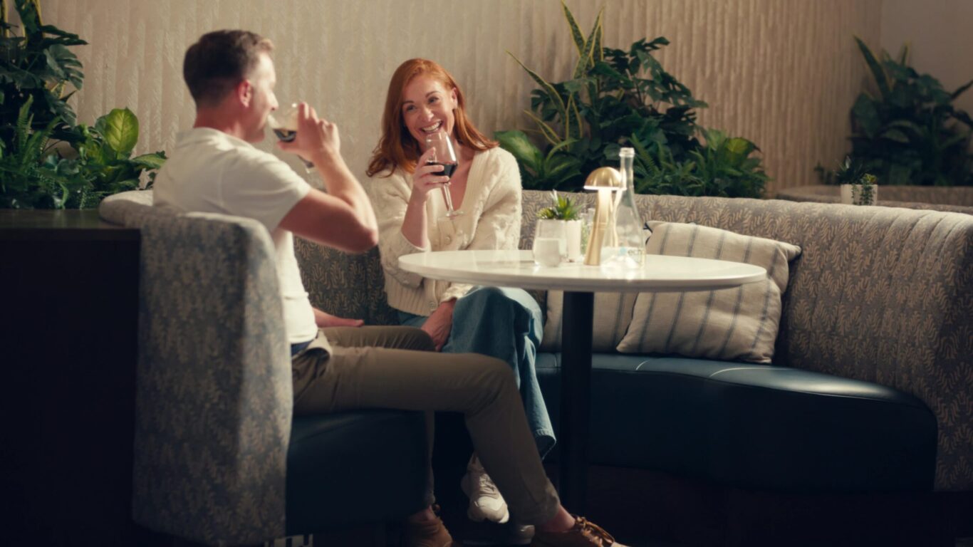 Two people sit at a round table in a modern lounge, smiling and drinking red wine, surrounded by plants and neutral decor.