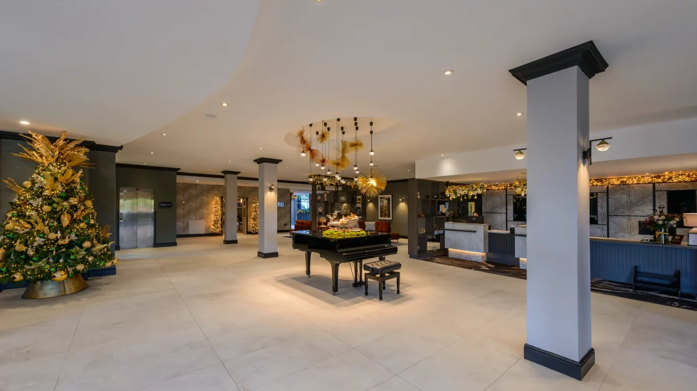 Spacious hotel lobby with a grand piano at the center, a decorated Christmas tree on the left, and a reception desk on the right.