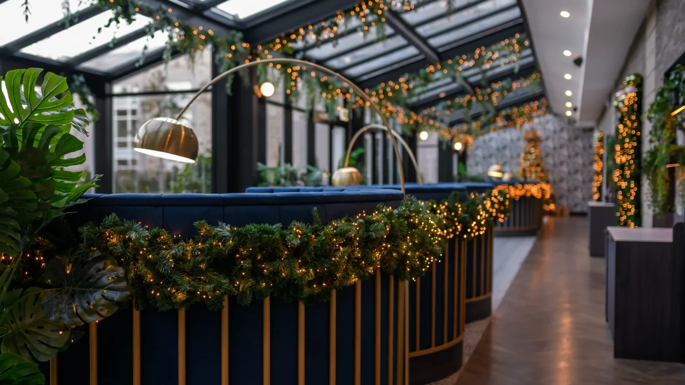 A modern indoor space with blue seating, gold lamps, and garlands decorated with string lights along a glass ceiling and walls.
