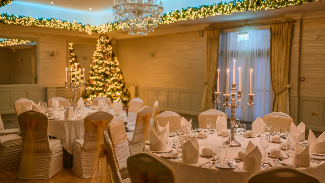 Banquet hall decorated for an event with round tables, white chair covers, candelabras, folded napkins, and Christmas lights, including a lit Christmas tree in the background.
