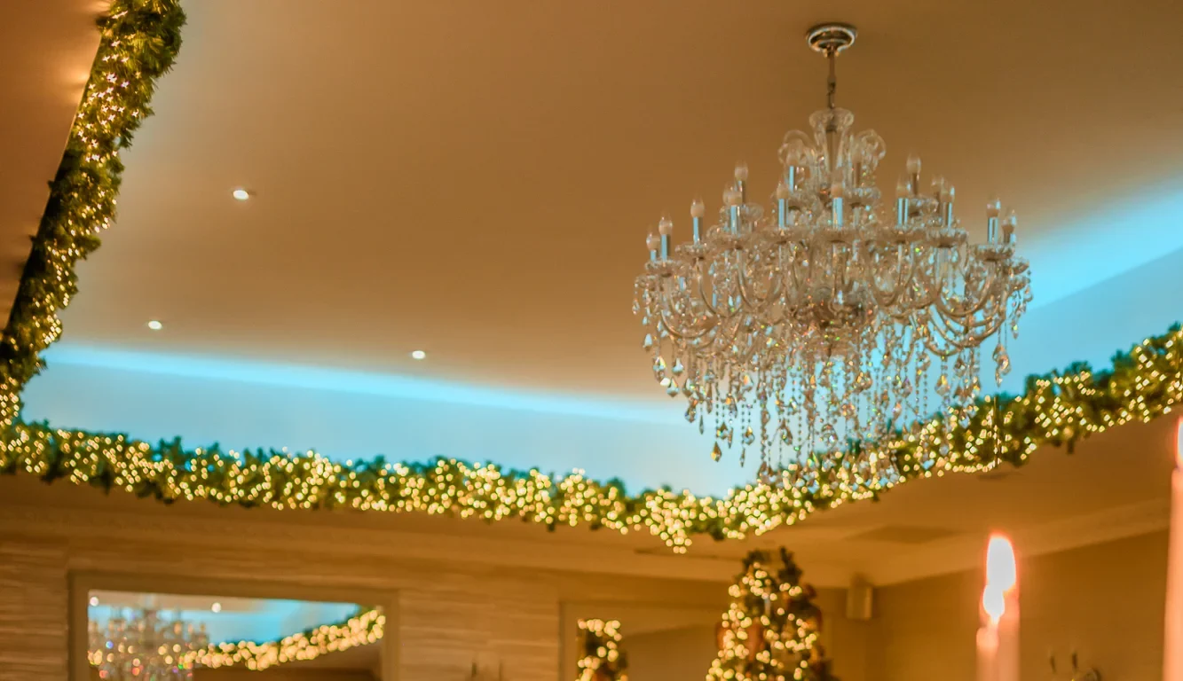 Elegant dining room set for an event, with round tables, white linens, candelabras, and decorated Christmas trees and garlands, under a crystal chandelier.