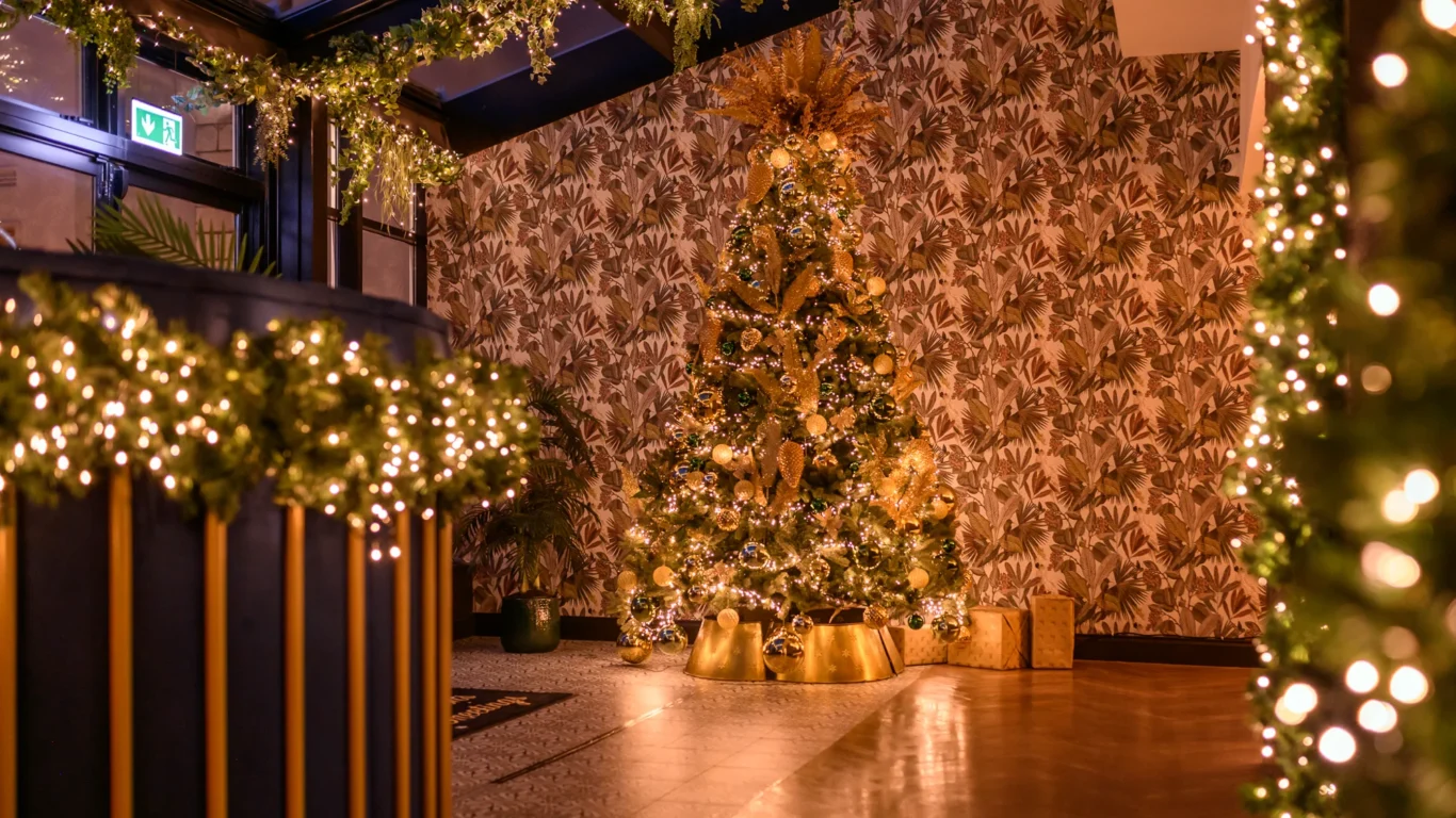 A decorated Christmas tree with gold ornaments and wrapped gifts stands in a warmly lit room with garlands and string lights.