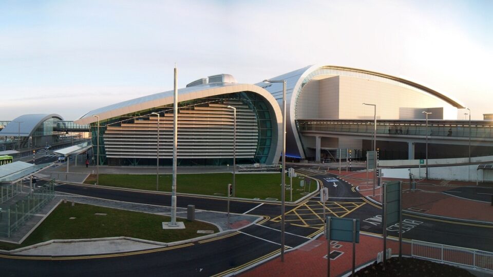 Modern airport terminal with curved glass and steel architecture, multiple roadways, bus stops, and elevated walkways visible under a clear sky.