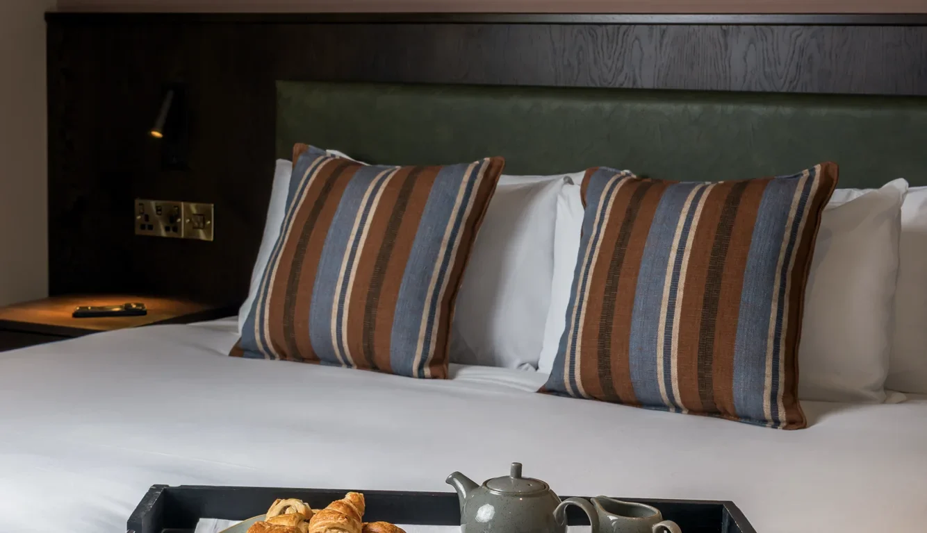 Elegant hotel room with striped cushions on bed, featuring a breakfast tray with pastries and tea for a relaxing stay.