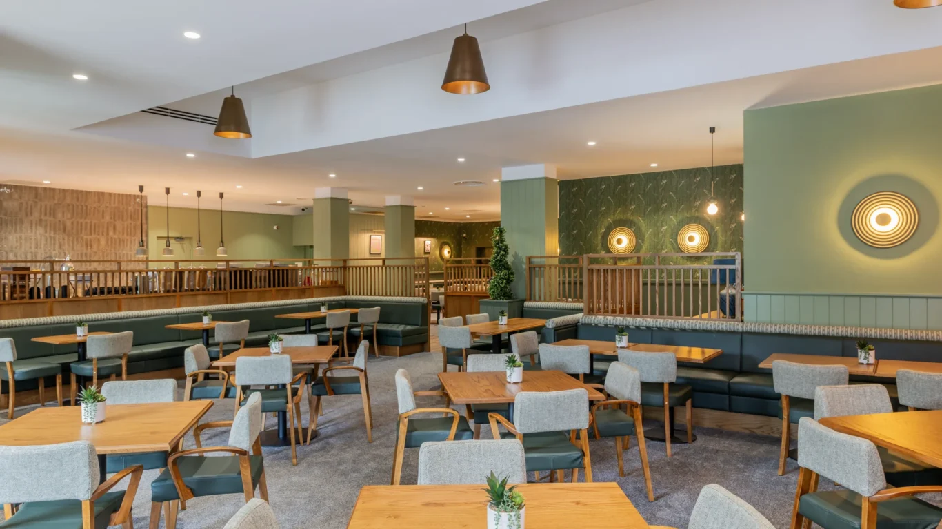 Modern hotel dining area with wooden tables, green accents, cosy seating, and warm lighting for a relaxing experience.
