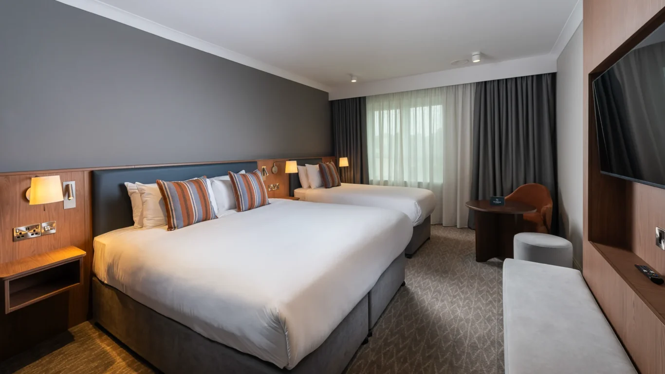 Elegant hotel room with two cosy beds, striped cushions, modern lighting, TV, and seating area, offering a comfortable stay.