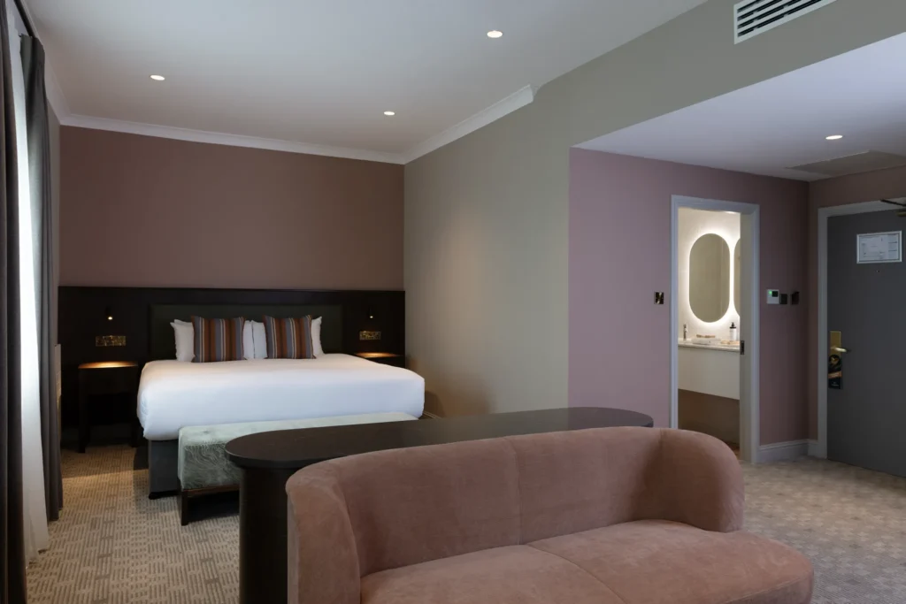 Elegant hotel room with a king-size bed, soft lighting, plush seating, and a modern en-suite bathroom. Cosy and inviting.