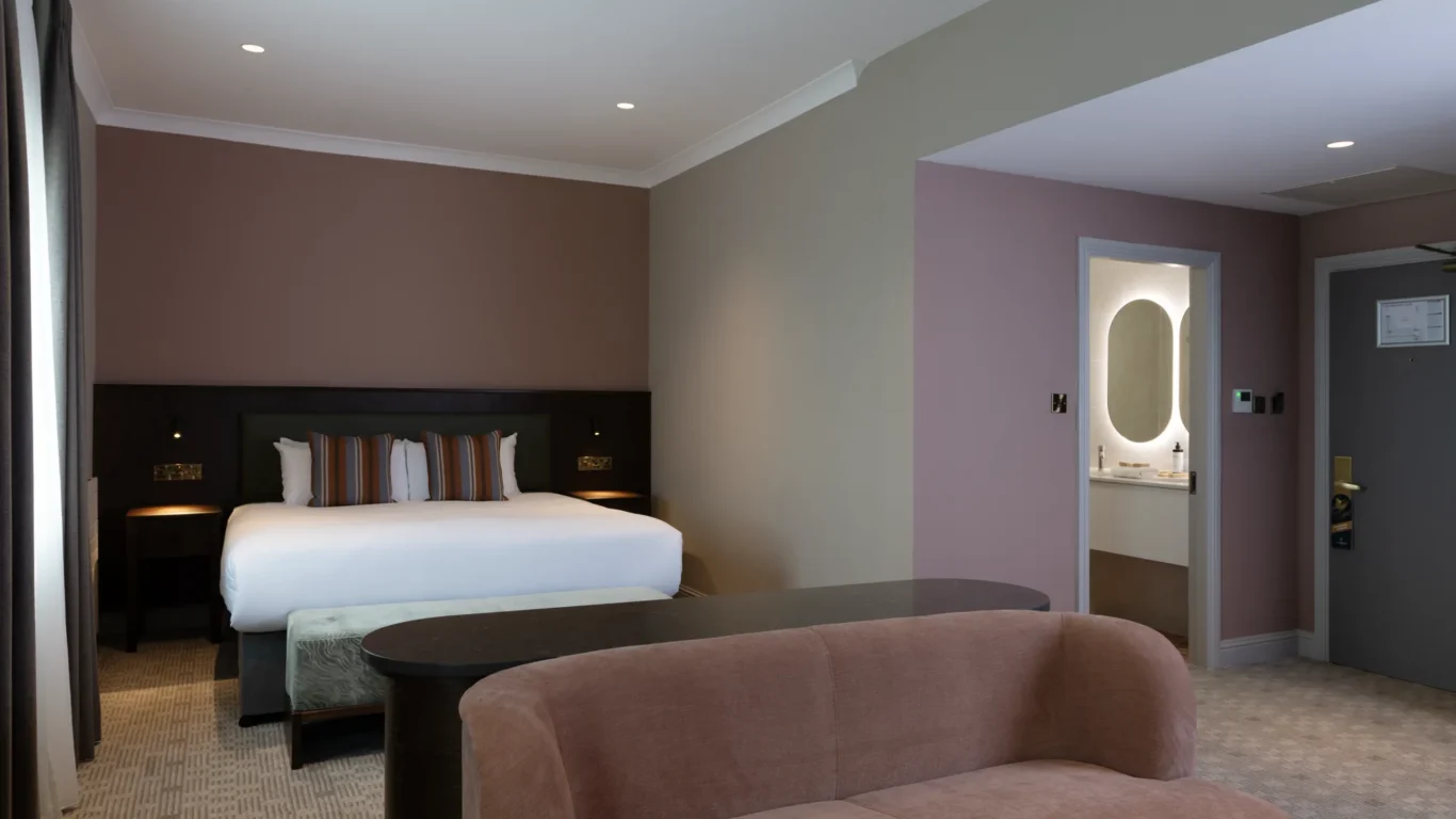 Elegant hotel room with a king-size bed, soft lighting, plush seating, and a modern en-suite bathroom. Cosy and inviting.
